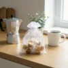Weißes Organzasäckchen 15x20 cm gefüllt mit Karamellbonbons auf einer Holzplatte neben einer Kaffeekanne.