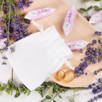 Weißer Säckchen für Gastgeschenke mit goldenen Eheringen, Lavendel und Süßigkeiten auf Holz.