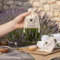 Hand hält Lavendelsäckchen mit Bienen-Motiv vor Lavendelfeld und Wellness-Zubehör